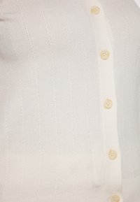 Prim-plan al unui cardigan tricotat de culoare crem, cu modele verticale și patru nasturi bej aberati în partea din față.