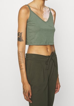 Slank person iført olivengrøn cropped tank top og matchende bukser med snøre, der viser vitiligo på skulderen og tatoveringer på arm og hånd.