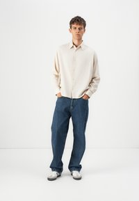 Cream button-up shirt with textured fabric, paired with loose blue denim jeans and white sneakers with gray accents. Model stands in a neutral background.