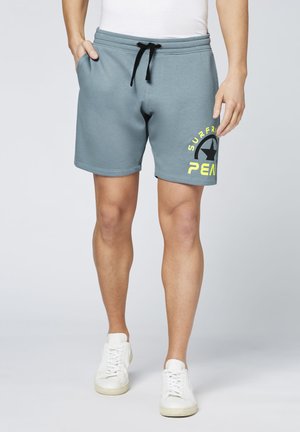 Graue Baumwollshorts mit einem schwarzen Kordelzug, seitlichen Taschen und gelbem Grafiktext am linken Bein. Kombiniert mit weißen Turnschuhen.