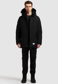 Juoda paminkštinta striukė su užtrauktuku, reguliuojama gaubtu ir keliais užtrauktukais užsegamais kišenėmis; derinama su juodomis karinių stiliaus kelnėmis ir batais.