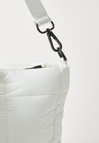 Sac à main blanc matelassé avec une texture lisse et moelleuse, doté d'un clip en métal détachable et d'une bandoulière tissée. Fermeture éclair sur le dessus.