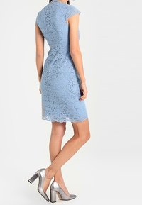 Hellblaues Spitzenkleid mit kurzen Ärmeln und betonter Taille. Silberne Schuhe mit Blockabsatz ergänzen das Outfit. Flaches Spitzenmuster sichtbar.