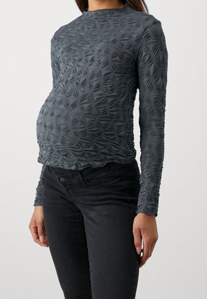 Femme portant un haut gris ajusté à manches longues avec un motif géométrique et un jean noir taille haute, debout devant un fond uni.