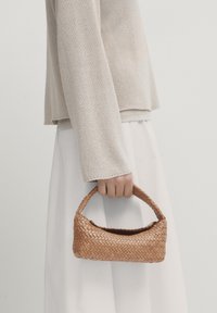 Main tenant un petit sac à main en osier marron, portant un pull en tricot beige et une jupe blanche devant un fond uni.