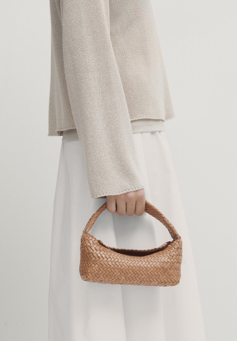 Main tenant un petit sac à main en osier marron, portant un pull en tricot beige et une jupe blanche devant un fond uni.