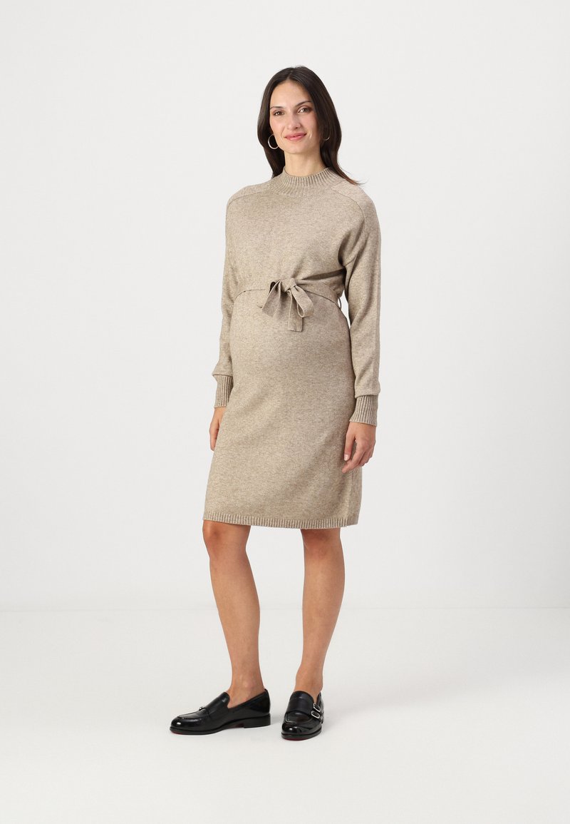 Abito maglione in beige, con collo rotondo, maniche lunghe e cintura in vita. Tessuto testurizzato con polsini e orlo a coste. Indossato con mocassini neri.