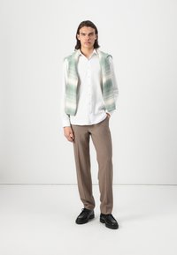 Chemise blanche à boutons, pull en plaid vert clair et crème drapé sur les épaules, pantalons beiges et chaussures noires. Fond uni.