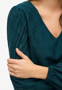 Teal blouse met een gestructureerd diamanten patroon, V-hals en pofmouwen met gathered manchetten. Close-up van de stof en een hand die de mouw vasthoudt.