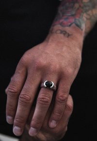 Sidabrinis žiedas su juodu akmeniu, turintis tekstūruotą juostą ir modernų, stilingą dizainą. Ranka rodo matomą odos tekstūrą ir tatuiruotę.