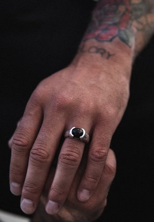 Silver ring with a black stone, featuring a textured band and a sleek, modern design. The hand shows visible skin texture and tattoo.
