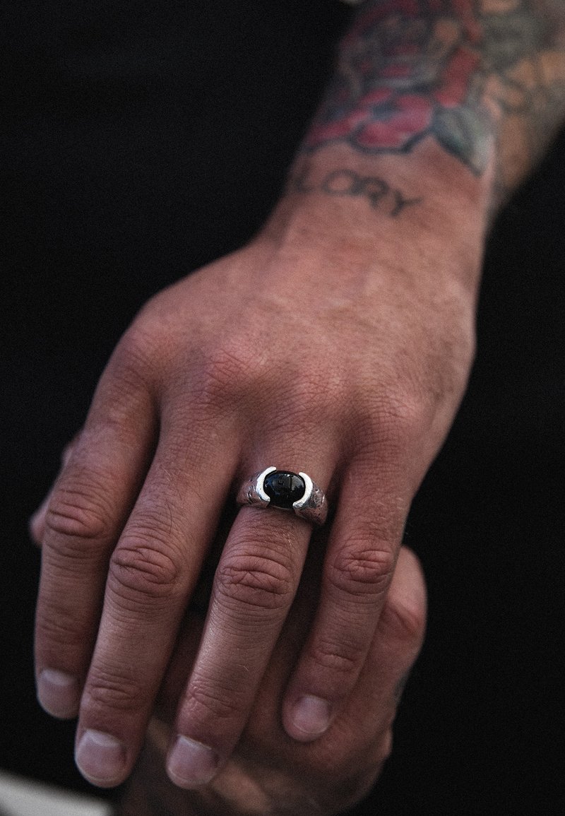 Sidabrinis žiedas su juodu akmeniu, turintis tekstūruotą juostą ir modernų, stilingą dizainą. Ranka rodo matomą odos tekstūrą ir tatuiruotę.