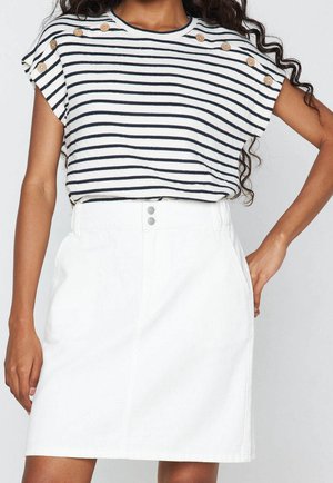 Femme portant un haut à manches courtes rayé avec des boutons en bois sur les épaules et une jupe blanche taille haute avec double rangée de boutons à l'avant.