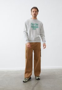 Sudadera gris con "Pier One Athletics 1968" en texto verde, combinada con pantalones cargo beige. El calzado presenta un patrón de camuflaje.