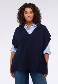 Granatowy dzianinowy ponczo z dekoltem w szpic, o luźnym fasonie i prążkowanej fakturze, noszony na jasnoniebieskiej koszuli na guziki.