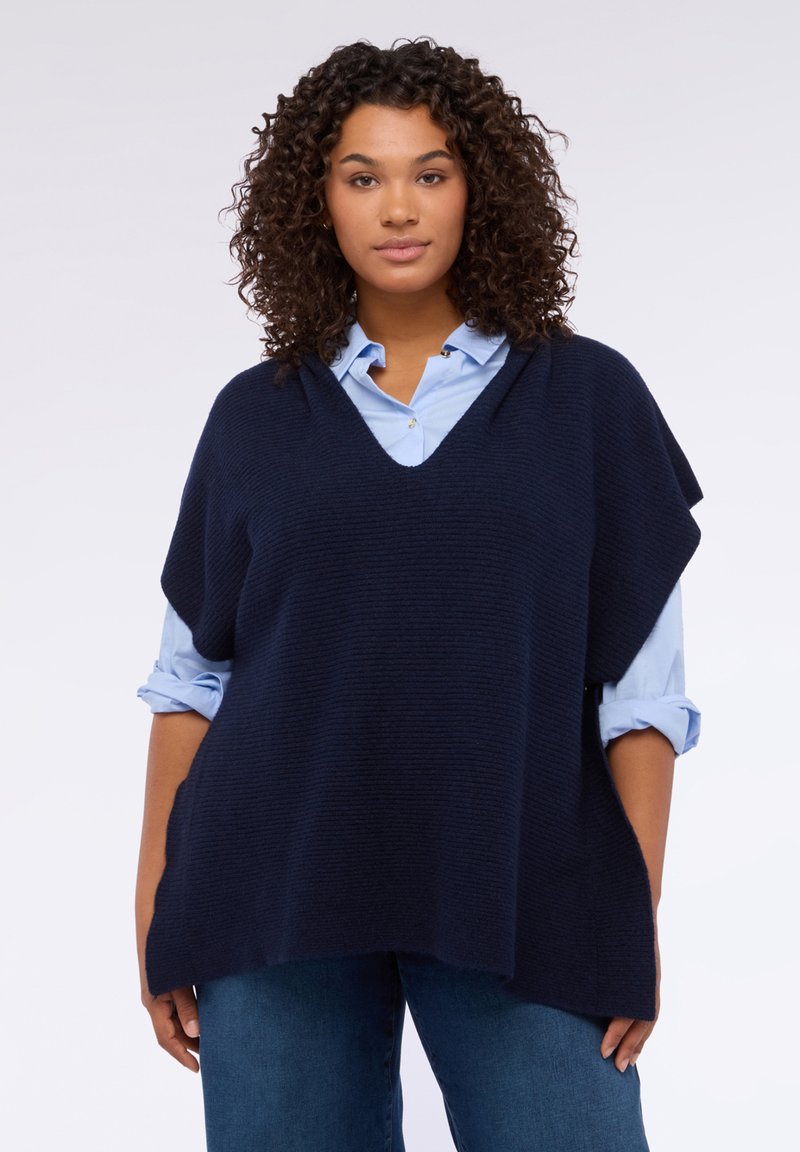 Granatowy dzianinowy ponczo z dekoltem w szpic, o luźnym fasonie i prążkowanej fakturze, noszony na jasnoniebieskiej koszuli na guziki.