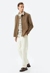 Brun denimjacka över en rutig skjorta, kombinerad med off-white jeans och mörka stövlar. Slät textur, knappdetaljer på jackan och skjortan.