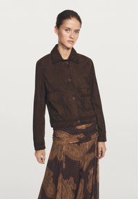 Bruine suède jas met een kraag, sluiting met knopen aan de voorkant en twee voorzakken. Het model draagt een gestreepte bruine rok.