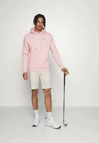 Svetloružová mikina s kapucňou a šnúrkami, v kombinácii s béžovými kraťasmi. Biely športový obuv s modrými akcentmi a golfová palica držaná v ruke.