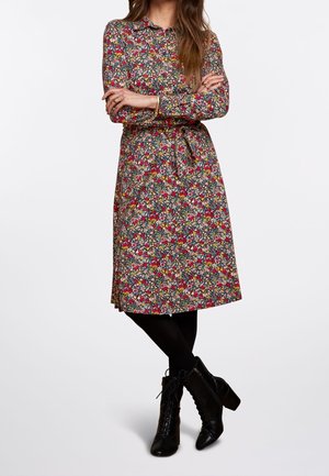 Femme portant une robe fleurie à manches longues avec un lien à la taille, des collants noirs et des bottines noires à talons lacées, debout, les bras croisés.