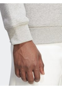 Šedá mikina s pružnými manžetami, strukturovanou tkaninou a detailním pohledem na ruku držící okraj. Jednoduchý a praktický design.