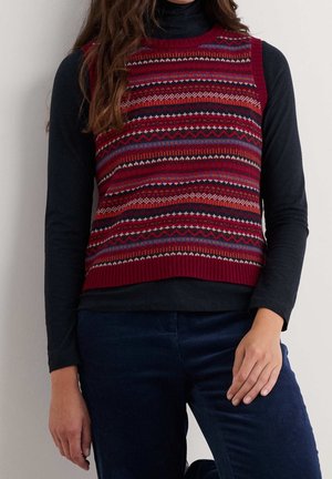Frau trägt eine ärmellose Strickweste mit roten, schwarzen und blauen geometrischen Mustern über einem schwarzen Langarmshirt und dunkelblauen Hosen.