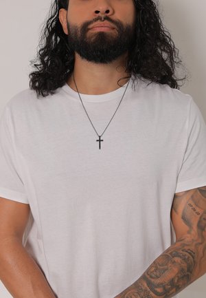 T-shirt blanc à col rond, manches courtes, porté avec une fine chaîne noire présentant un pendentif en croix. Tatouage visible sur l'avant-bras.