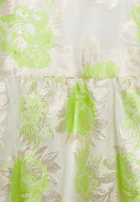 Genomskinligt tyg med ett blommönster i klara gröna och gyllene toner, med en mjuk textur och subtil lyster. Rynkdetalj vid midjan.