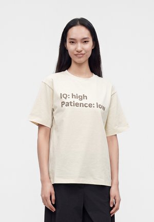 Joven mujer con cabello largo y oscuro lleva una camiseta beige con texto "IQ: high Patience: low" y pantalones negros sobre un fondo liso.