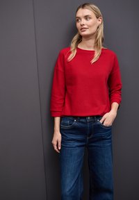 Maglione rosso a coste con maniche 3/4, abbinato a jeans a gamba larga blu scuro. Design semplice senza motivi o accenti visibili.