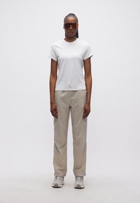 Γυναίκα που στέκεται, φοράει λευκό μπλουζάκι, μπεζ παντελόνι cargo, γκρι αθλητικά παπούτσια και μεγάλα πορτοκαλί γυαλιά ηλίου, σε λευκό φόντο.