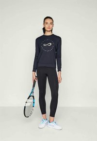 Sötétkék, hosszú ujjú pulóver kör alakú logóval, fekete leggings-szel párosítva. A modell egy kék és fekete teniszütőt tart a kezében, fehér sportcipőt visel.