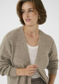 Femme aux cheveux bruns courts portant de grandes boucles d'oreilles en or, un cardigan beige par-dessus un débardeur blanc à côtes, tenant l'avant du cardigan, regardant vers la droite.
