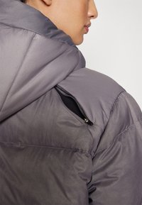 Veste rembourrée à capuche en tissu gris avec une texture lisse ; détail de poche zippée sur le côté visible sur l'épaule gauche.