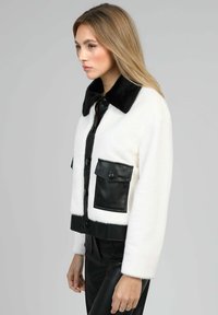 Veste noire et blanche cropped avec une texture en fourrure, dotée d'un col noir, de boutons à l'avant et de deux poches en cuir noir.