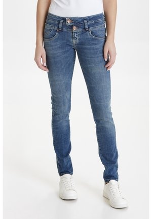 Kobieta nosząca niebieskie jeansy slim fit z podwójnymi guzikami w pasie i białe tenisówki, stojąca na jednolitym białym tle.