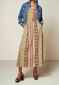 Robe longue en tissu à motifs avec des designs jaunes, rouges et marron, associée à une veste en jean bleue et des sandales marron à talons blocs.