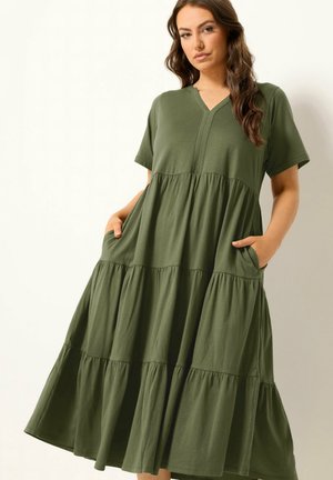 Kobieta ubrana w luźną, warstwową oliwkowozieloną sukienkę z krótkimi rękawami, dekoltem w szpic i rękami w kieszeniach, stojąca na jasnym, jednolitym tle.