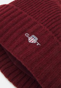 Bordeaux gebreide beanie met ribbeltextuur en een omgevouwen rand. Voorzien van een geborduurd embleem met rode, blauwe en witte accenten.