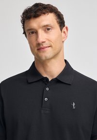 Svart polo-tröja med klassisk krage, treknappslå, strukturerad tyg och en liten broderad logotyp på vänster bröst.