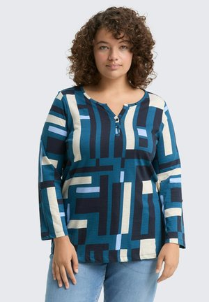 Femme aux cheveux bouclés portant un haut à manches longues à motifs géométriques bleu, noir et beige, avec un jean bleu clair, se tenant contre un fond uni.