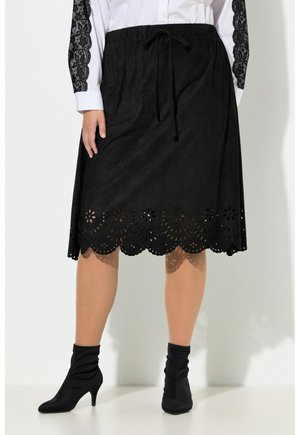 Czarna spódnica do kolan z falistym, laserowo wycinanym kwiatowym dołem, zestawiona z czarnymi botkami na obcasie i białą bluzką z czarnymi koronkowymi rękawami.