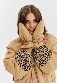 Žena v béžovej nafúkanej bunde s ružovými detailmi a leopardími lakťovými nášivkami, drží béžové rukavice pri lícach a usmieva sa.