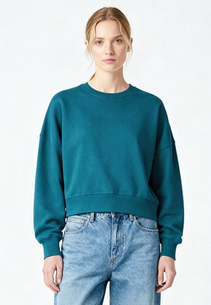 Teal cropped sweatshirt in soft fabric, featuring a round neckline and dropped shoulders. Paired with high-waisted light blue jeans.