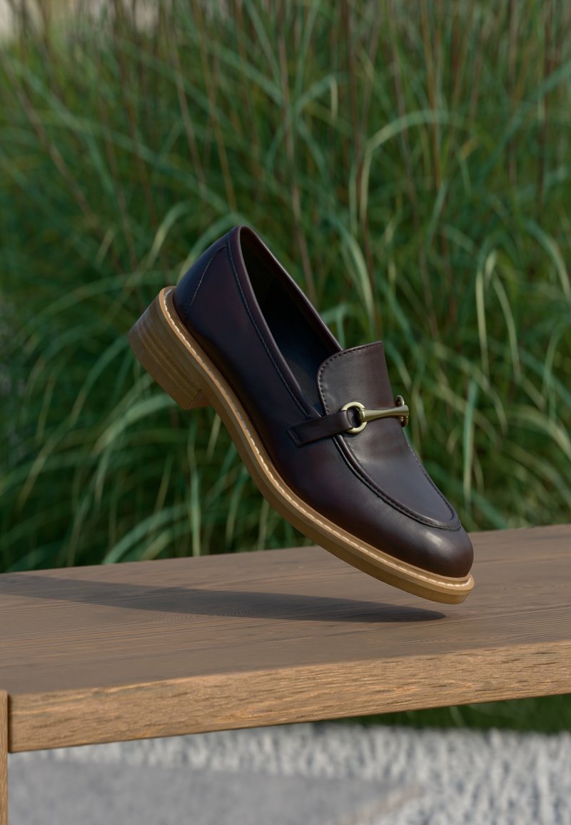 Mocassin en cuir brun foncé avec semelle en bois et détail en métal doré, suspendu au-dessus d'un banc en bois avec un fond d'herbe verte.