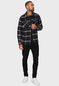 Camisa de franela a cuadros en blanco y negro con detalles en naranja, dos bolsillos en el pecho, cierre de botones, llevada sobre una camiseta negra y combinada con pantalones negros y zapatillas blancas.