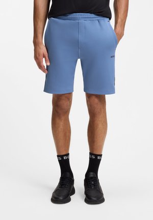 Hombre con pantalones cortos azul claro hasta la rodilla, calcetines negros con texto blanco "BOSS" y zapatillas negras, de pie con las manos en los bolsillos.