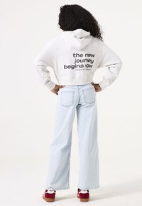 Grijze cropped hoodie met "de nieuwe reis begint nu" gedrukt op de achterkant, gecombineerd met lichtblauwe wijde jeans en rood-witte sneakers.
