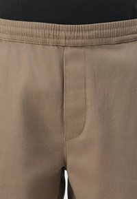 Nahaufnahme von beigen Hosen mit elastischem Bund und vorderer Naht, getragen von einer Person mit schwarzem Oberteil.