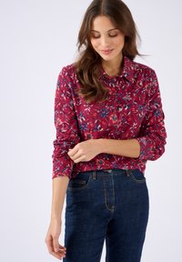 Donna con lunghi capelli castani che indossa una camicia a fiori rossa con bottoni e jeans di un blu scuro, che sistema la manica sinistra con un leggero sorriso.